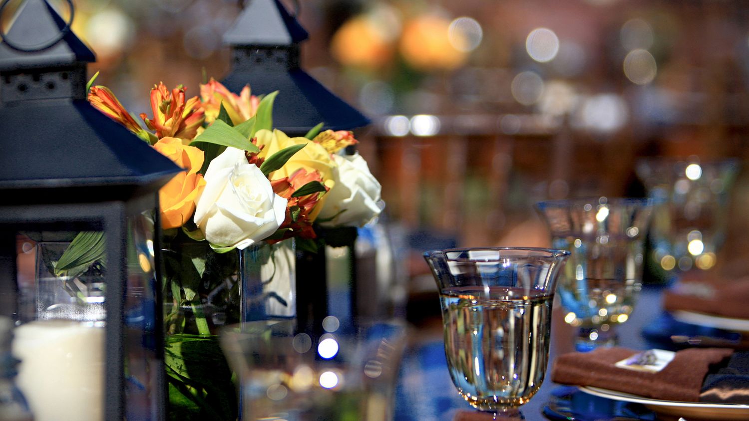 Elegant event table setting with blue linens, lanterns, orange and white floral centerpiece, and crystal glassware.