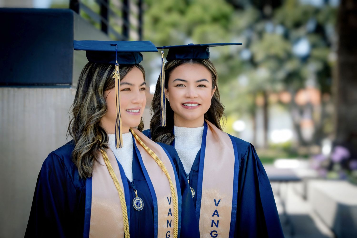 Two smiling women in blue graduation caps and gowns with gold stoles pose outdoors on a sunny campus day.