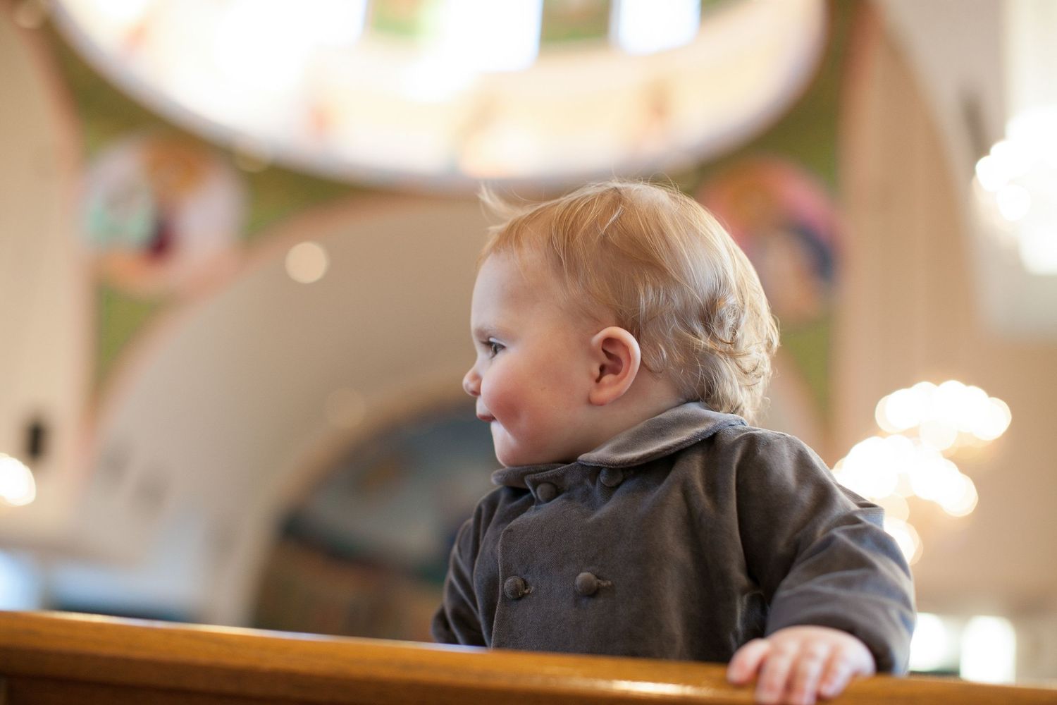 Adorable baby in gray jacket standing at wooden railing, smiling in bright indoor setting.