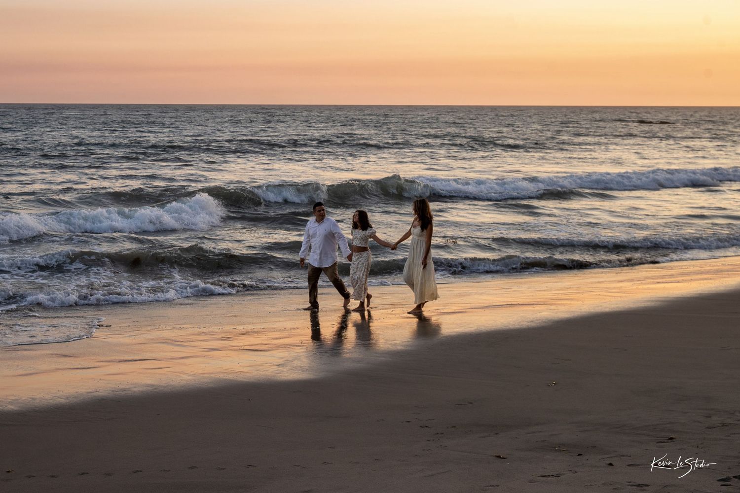 Family of three holding hands walking on a sandy beach at sunset with gentle ocean waves in the background.