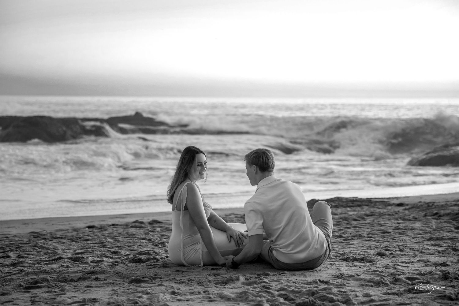 Black and white photo of a couple sitting on a sandy beach, facing each other with ocean waves in the background.