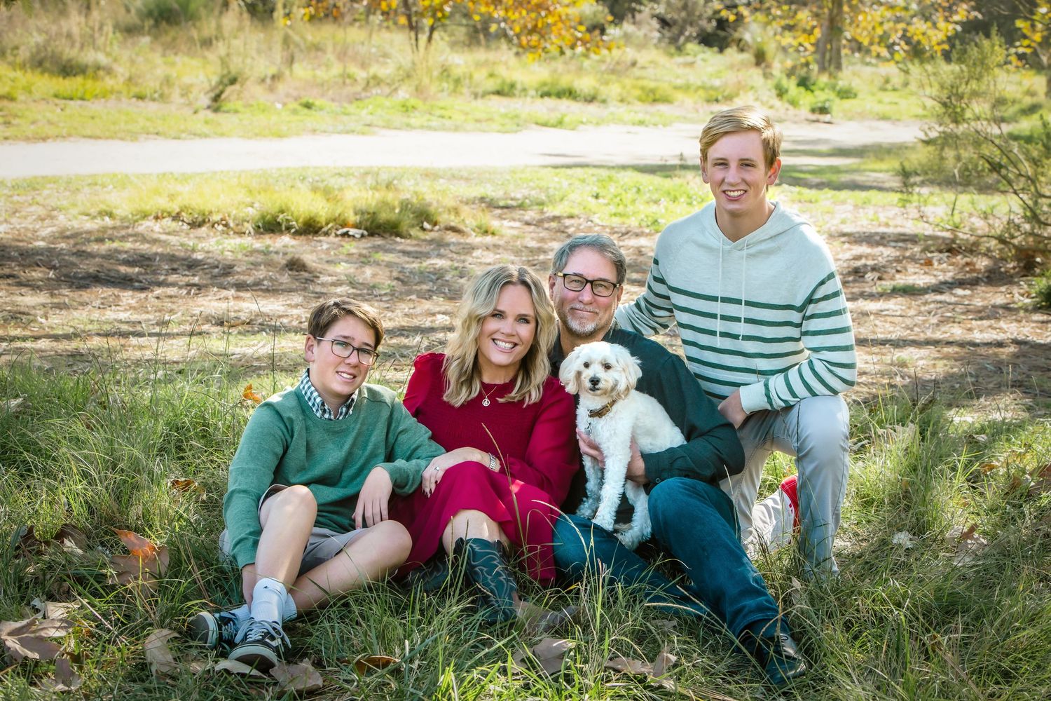 Family of four sits in green grass with their white dog in a sunny outdoor portrait.