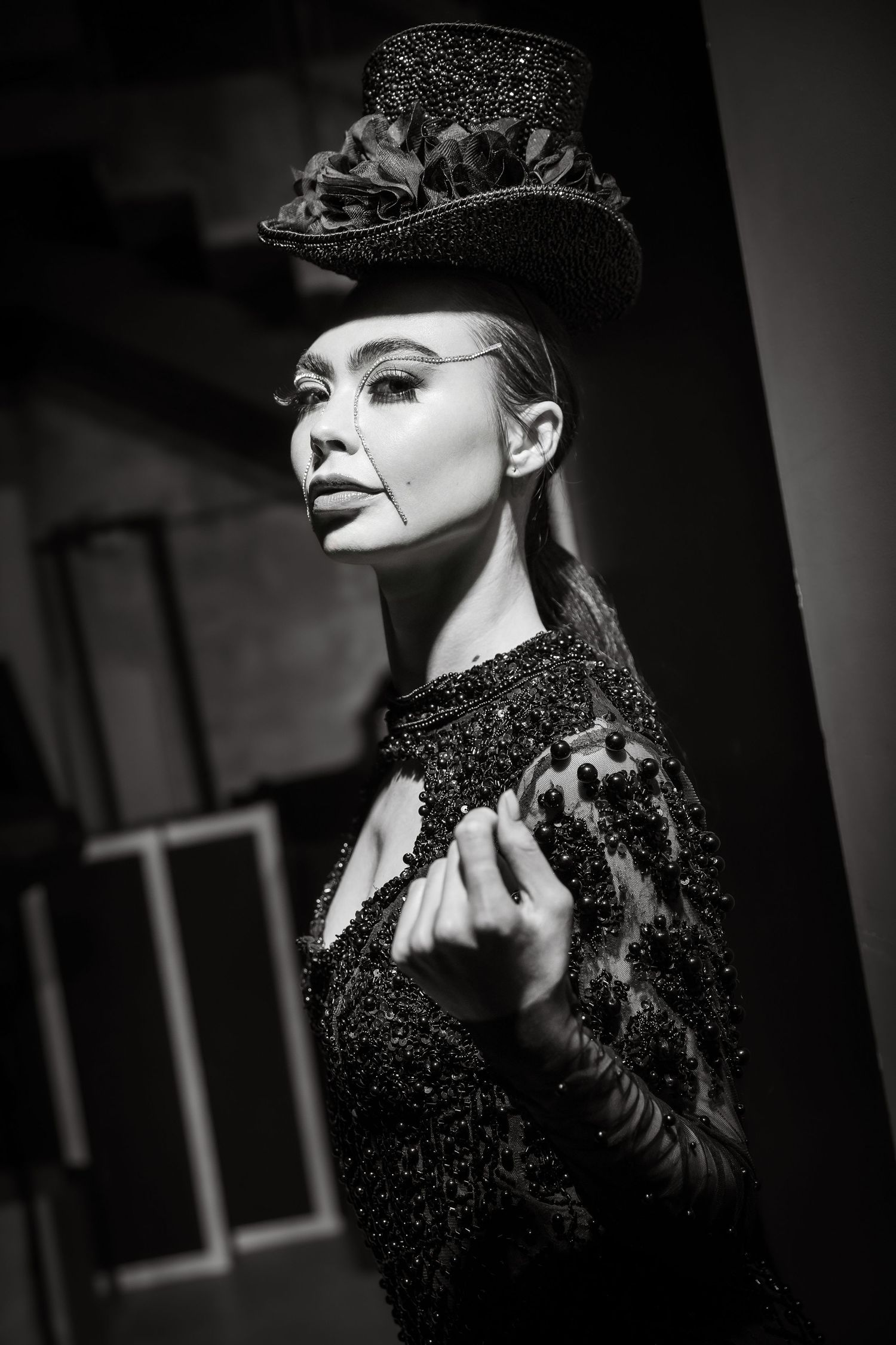 Black and white portrait of a model in dramatic makeup wearing a sparkly black top hat and embellished dark gown.
