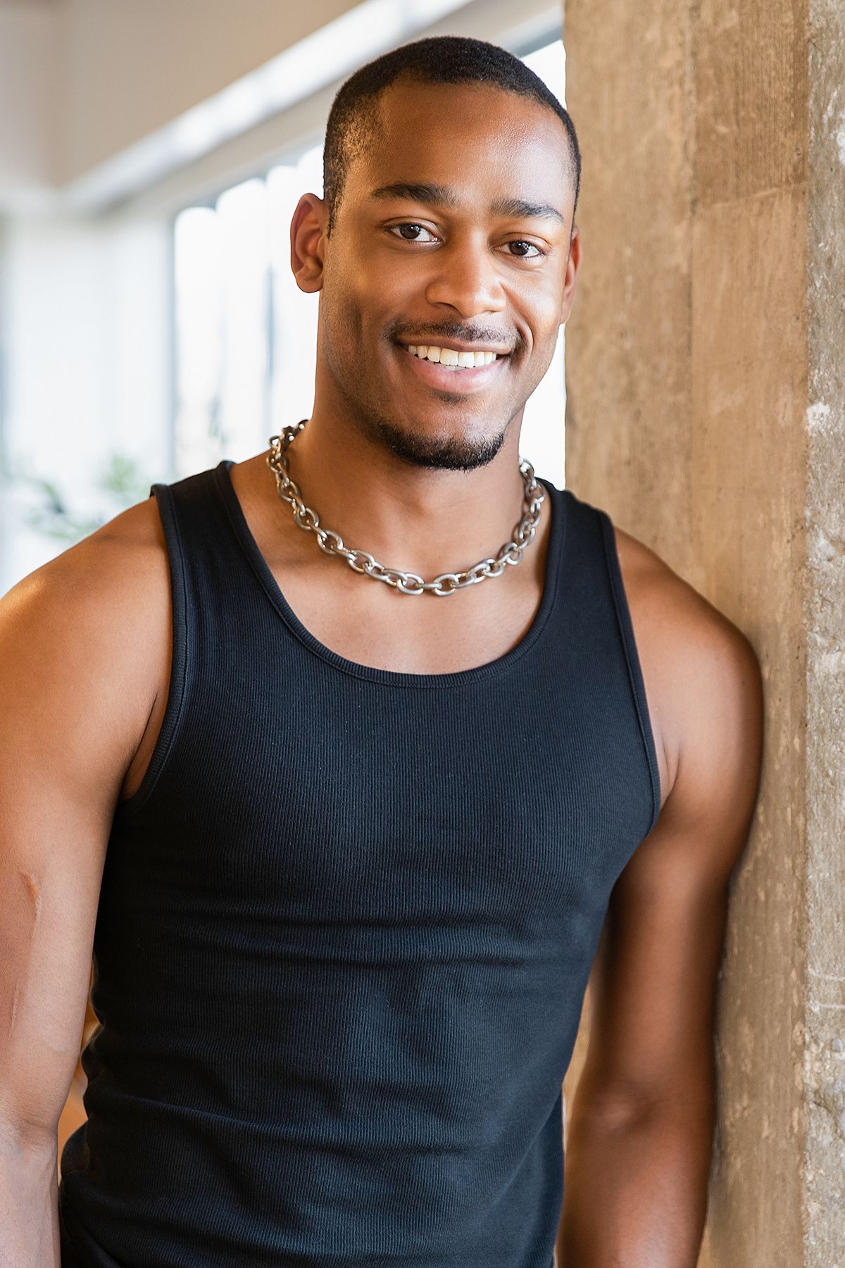 Smiling young Black man with short hair wearing a black tank top and chain necklace, leaning against a wooden pillar.