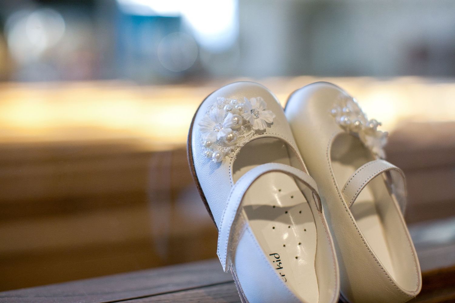 White flower girl shoes with floral embellishments resting on a wooden church pew.