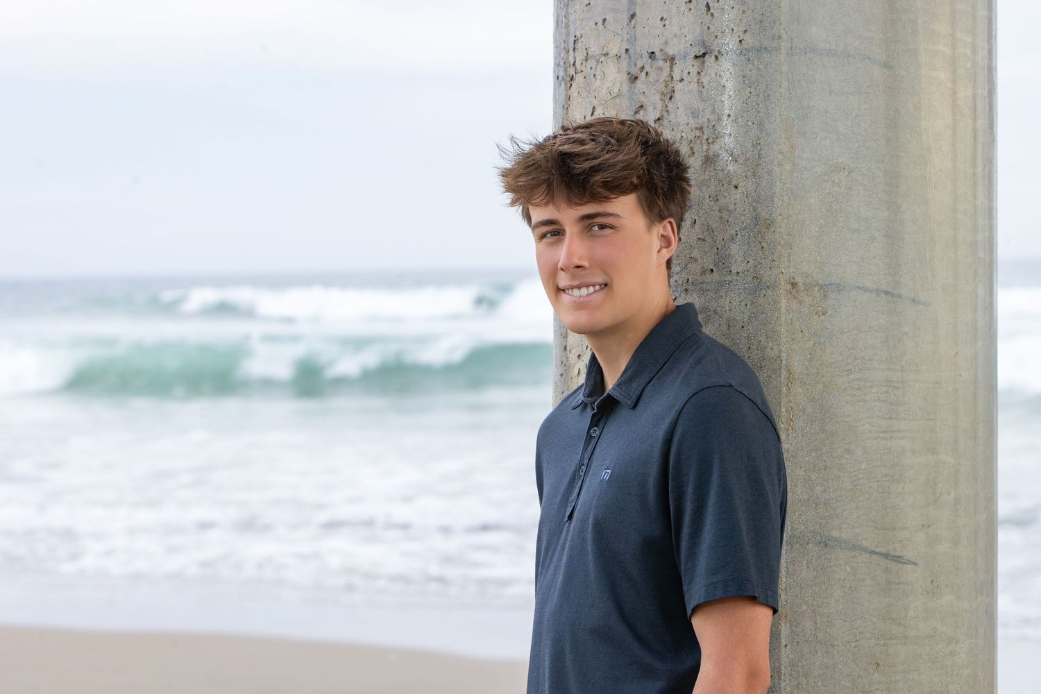 Young man in navy polo shirt leaning against a concrete pier pillar on a sandy beach with ocean waves behind him.