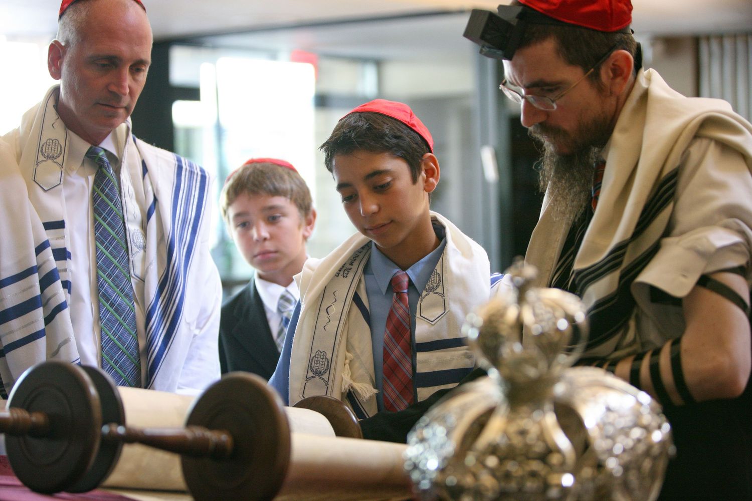 Jewish boy reads Torah scroll during Bar Mitzvah ceremony with rabbi and family wearing tallit prayer shawls.