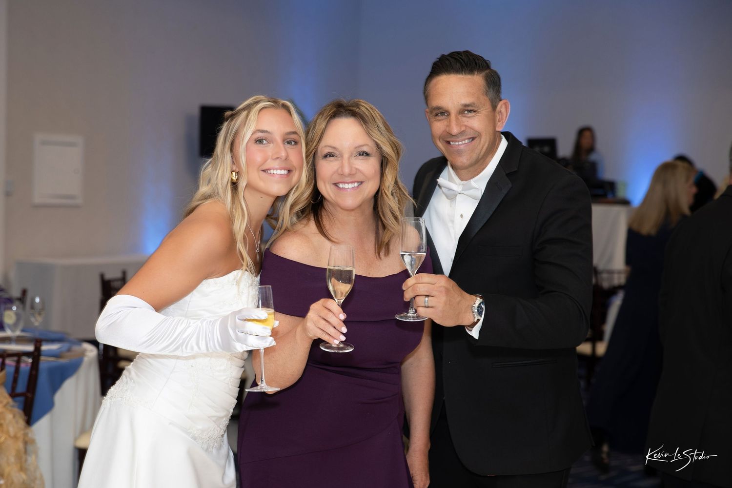Three guests smile and hold champagne glasses at an elegant wedding reception in a bright venue.