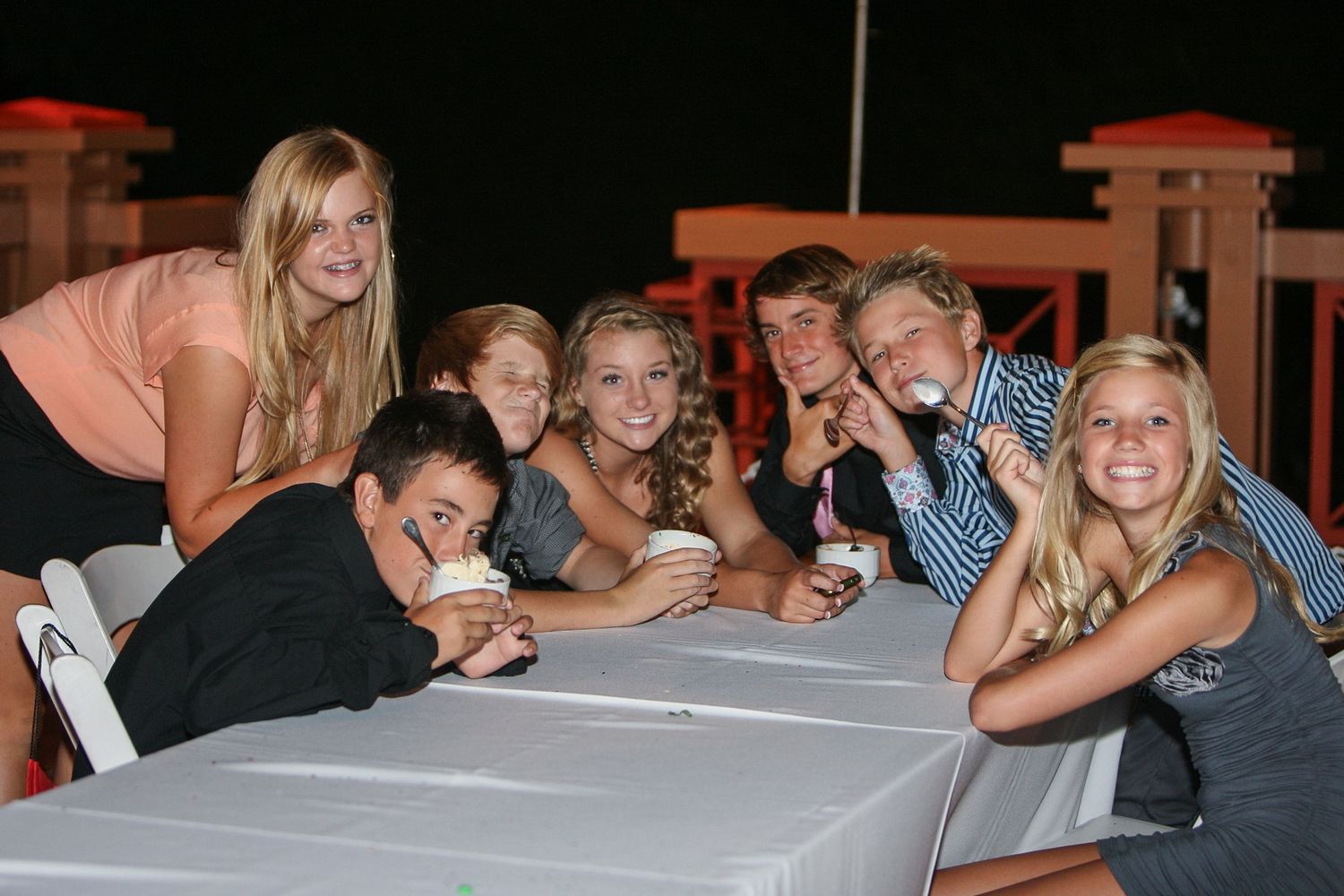 Group of teenagers smiling and posing at an outdoor table at night, holding desserts and enjoying a summer gathering.
