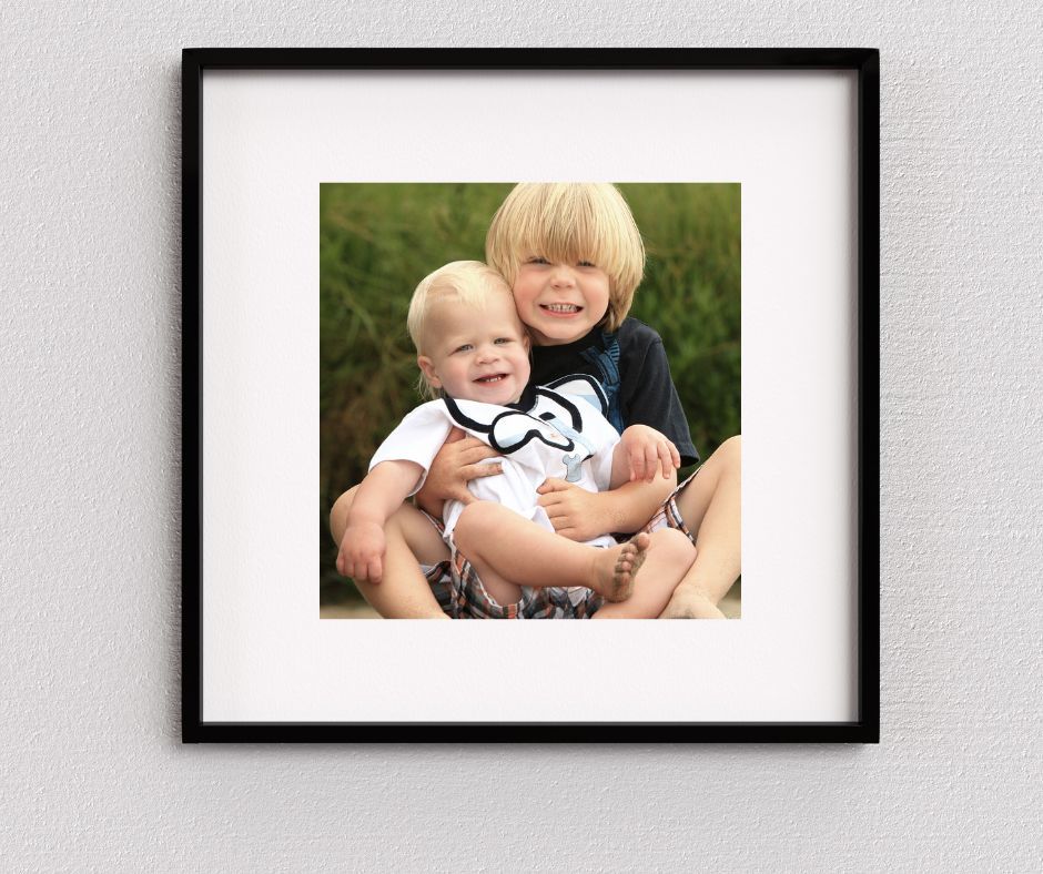 Framed photo of two young brothers, older blonde boy hugging smiling baby wearing mustache onesie, sitting outdoors.
