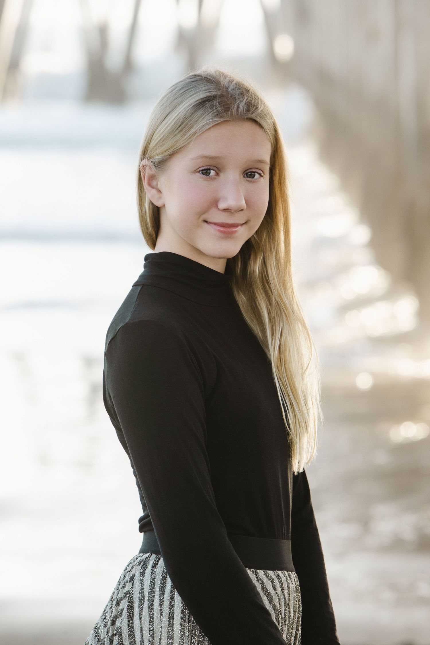 A young blonde girl in a black turtleneck and striped skirt smiles softly outdoors in a winter setting.