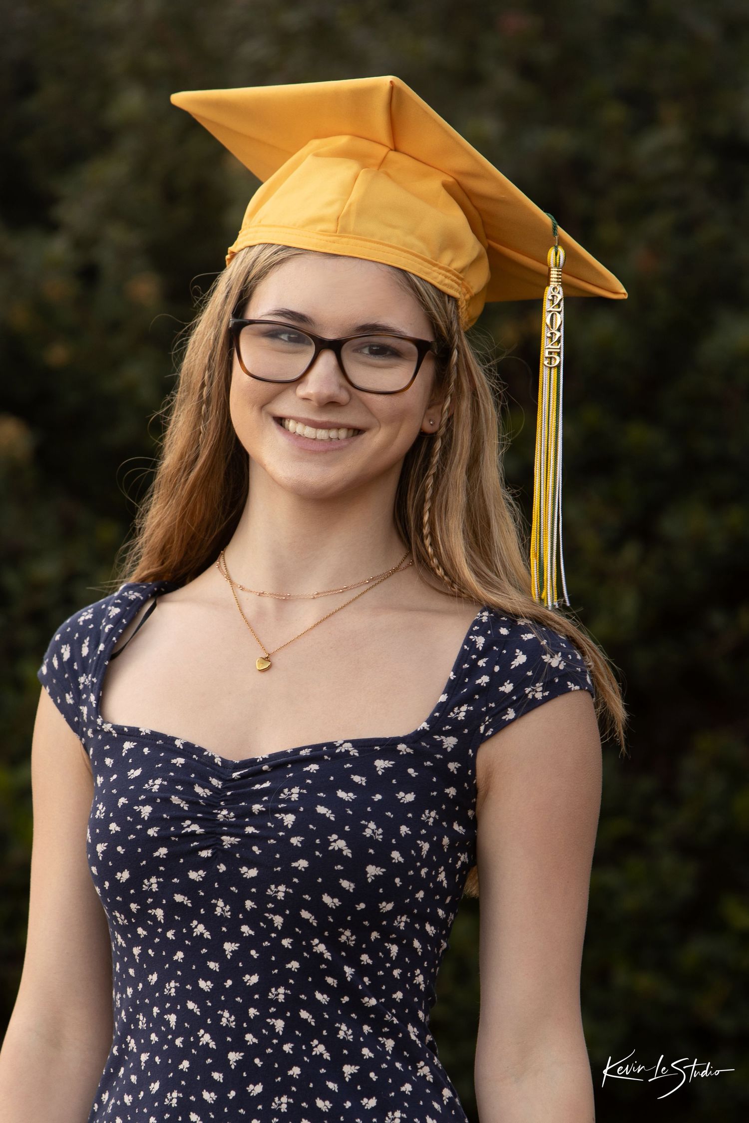 Smiling graduate in gold cap and navy floral dress poses outdoors for senior portrait.