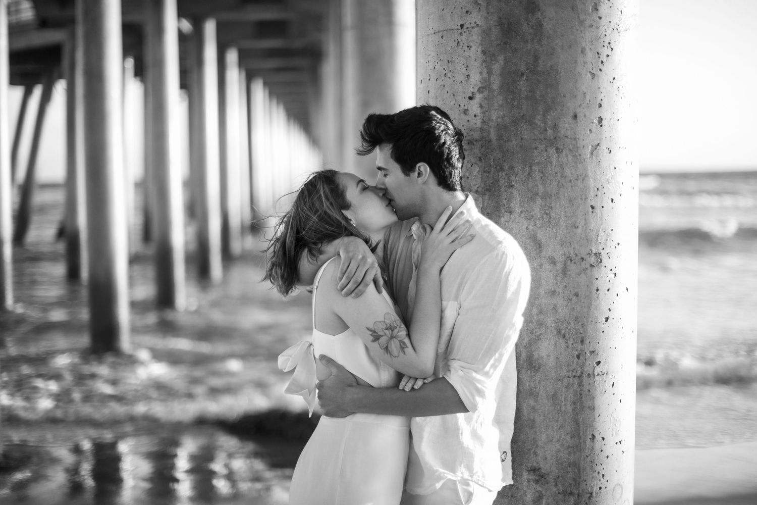 Black and white photo of a couple sharing a passionate kiss under a seaside pier.