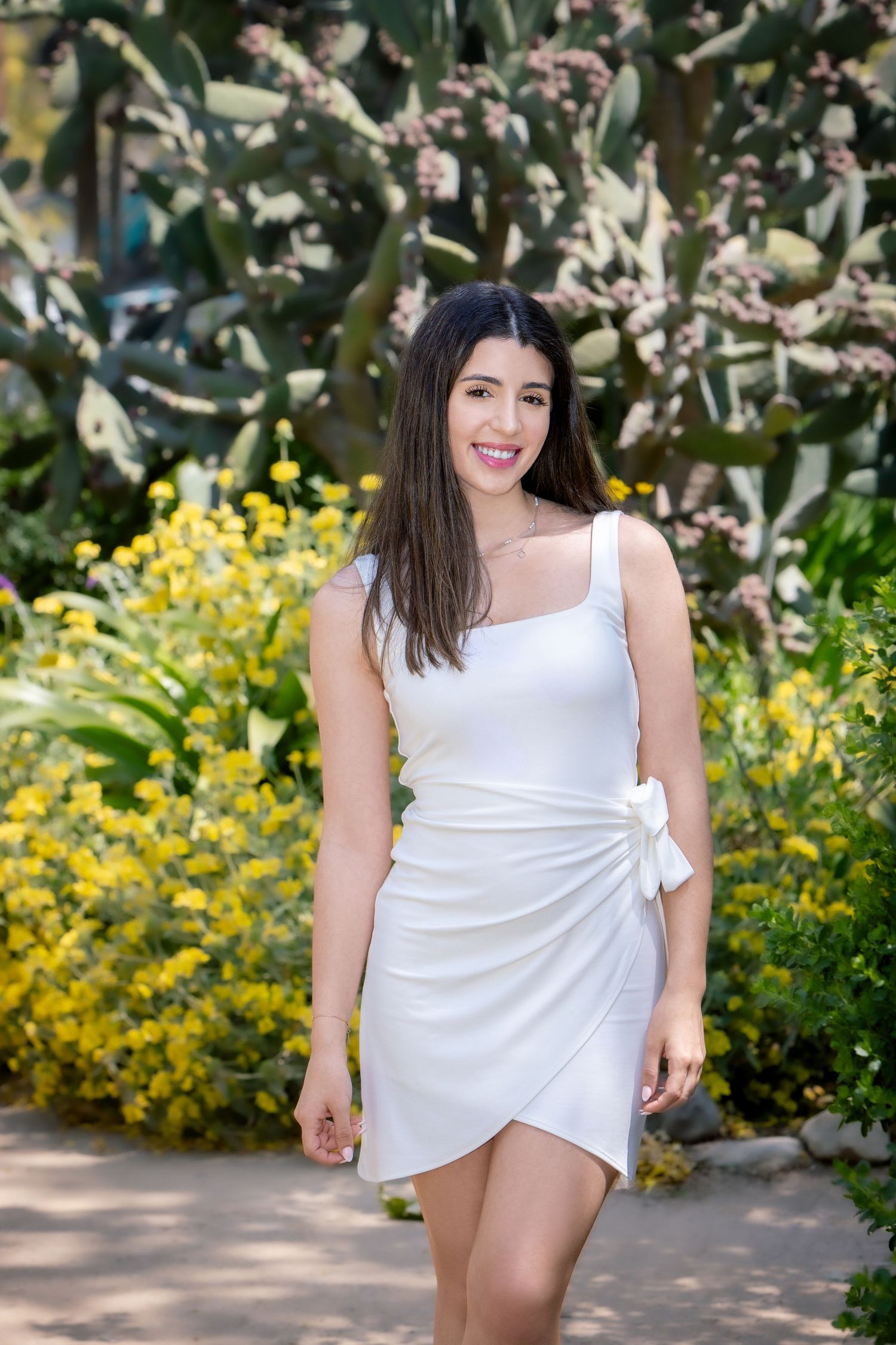 Smiling young woman in white wrap mini dress poses among yellow flowers and cacti outdoors.