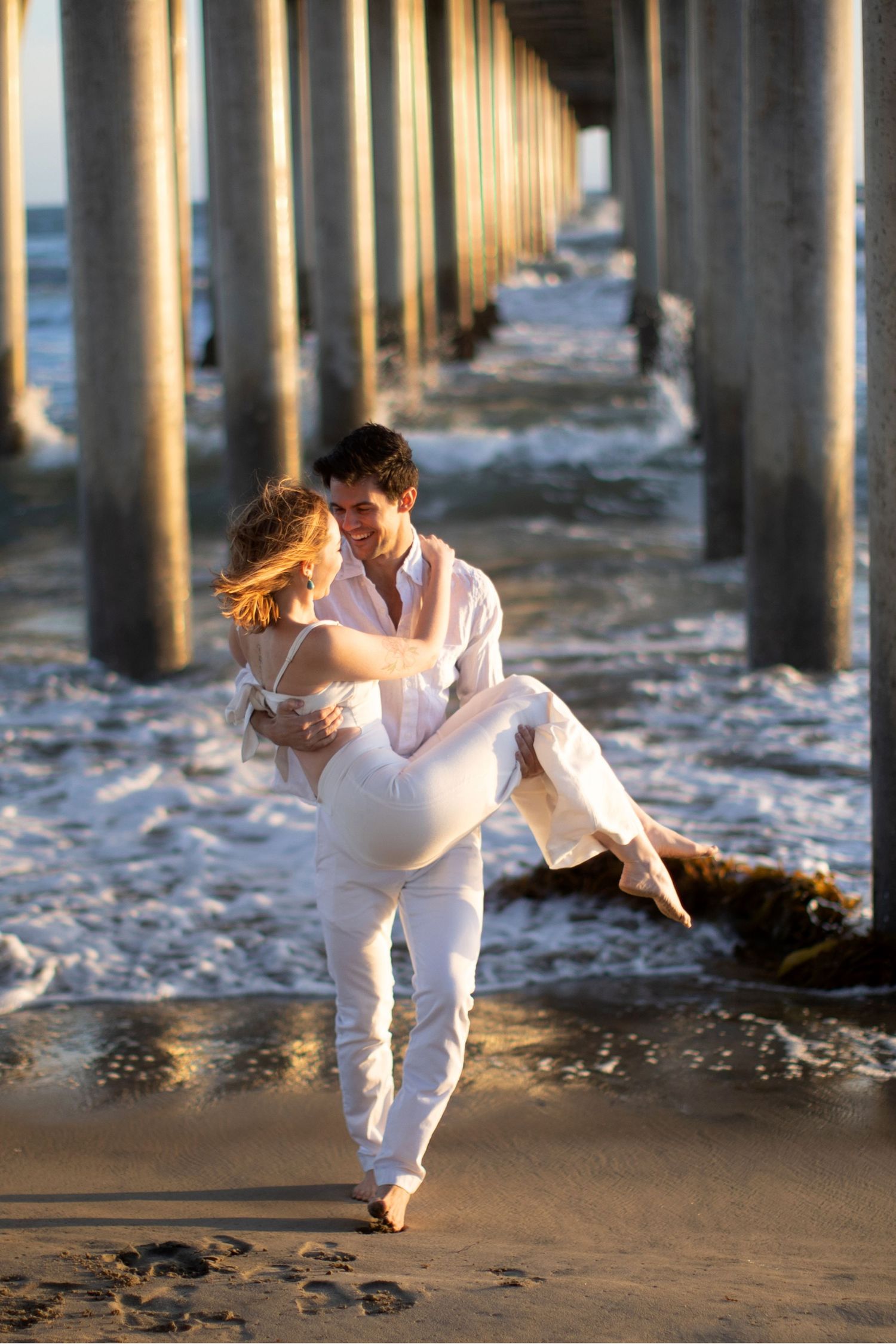 Man carrying woman in white outfits under a sunlit pier with ocean waves at sunset.