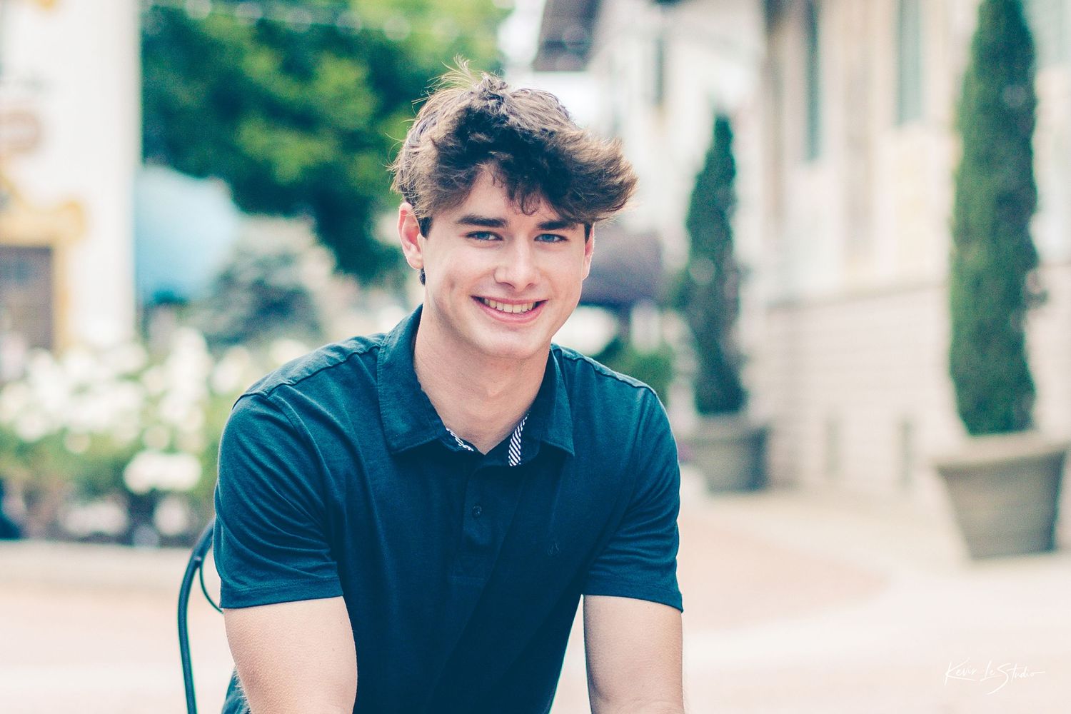Smiling young man in teal polo shirt seated outdoors in a bright, tree-lined village courtyard.