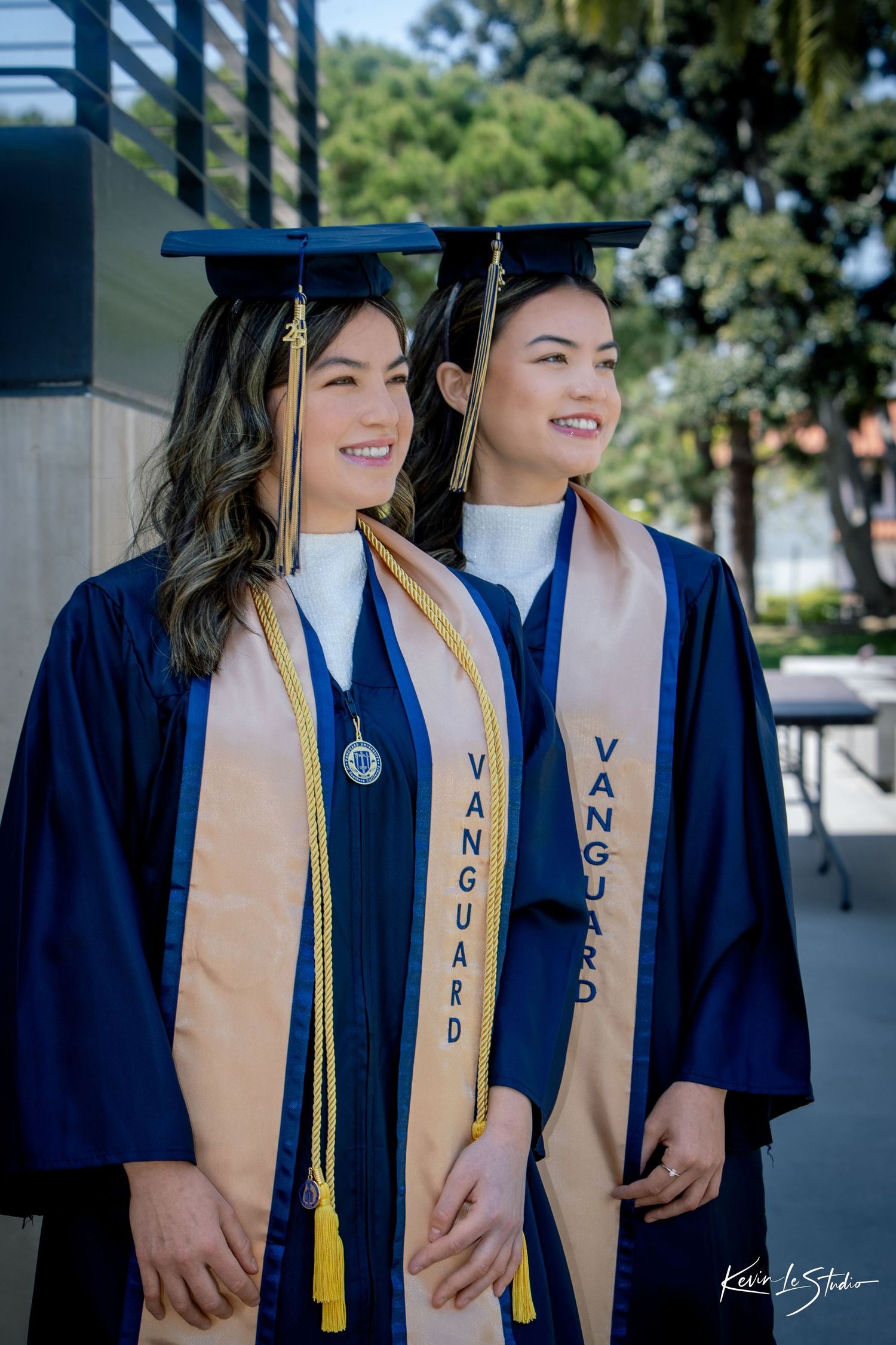 Two smiling graduates in navy Vanguard University caps and gowns pose together outdoors.