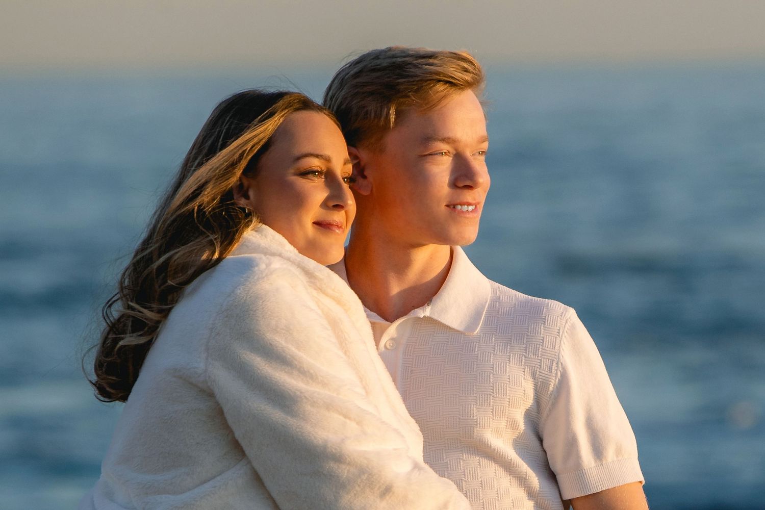 Couple embracing at golden hour by the ocean, woman in white wrap and man in white polo shirt.