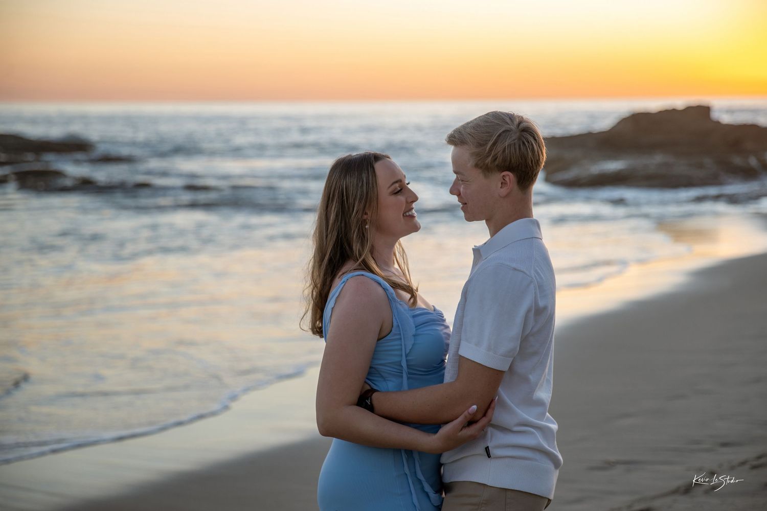 Young couple facing each other smiling on sandy beach during vibrant orange sunset.