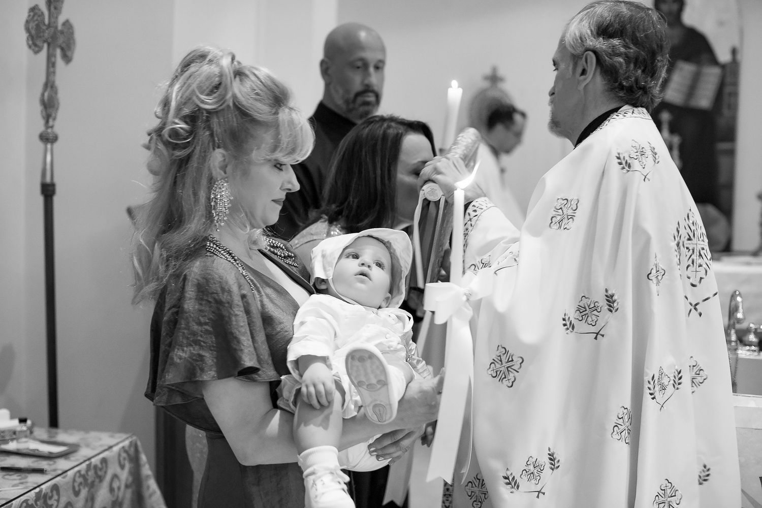 Black and white photo of a baby baptism ceremony with a priest, mother holding infant in white christening gown.