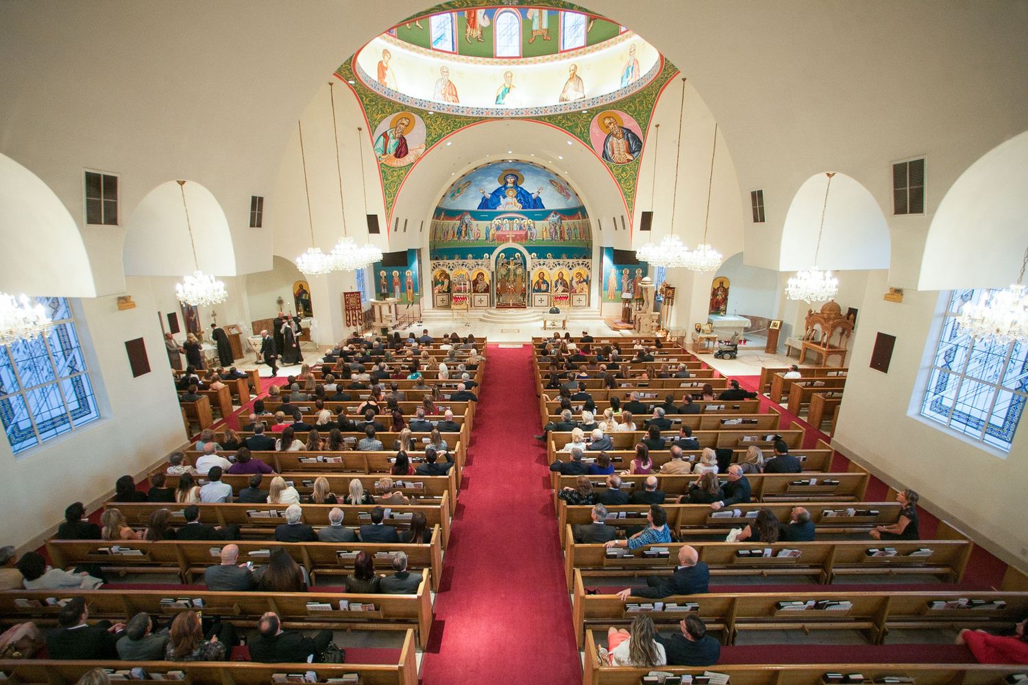 Crowded church interior with wooden pews, red carpet aisle, and ornate Byzantine mosaic altar during a religious service.