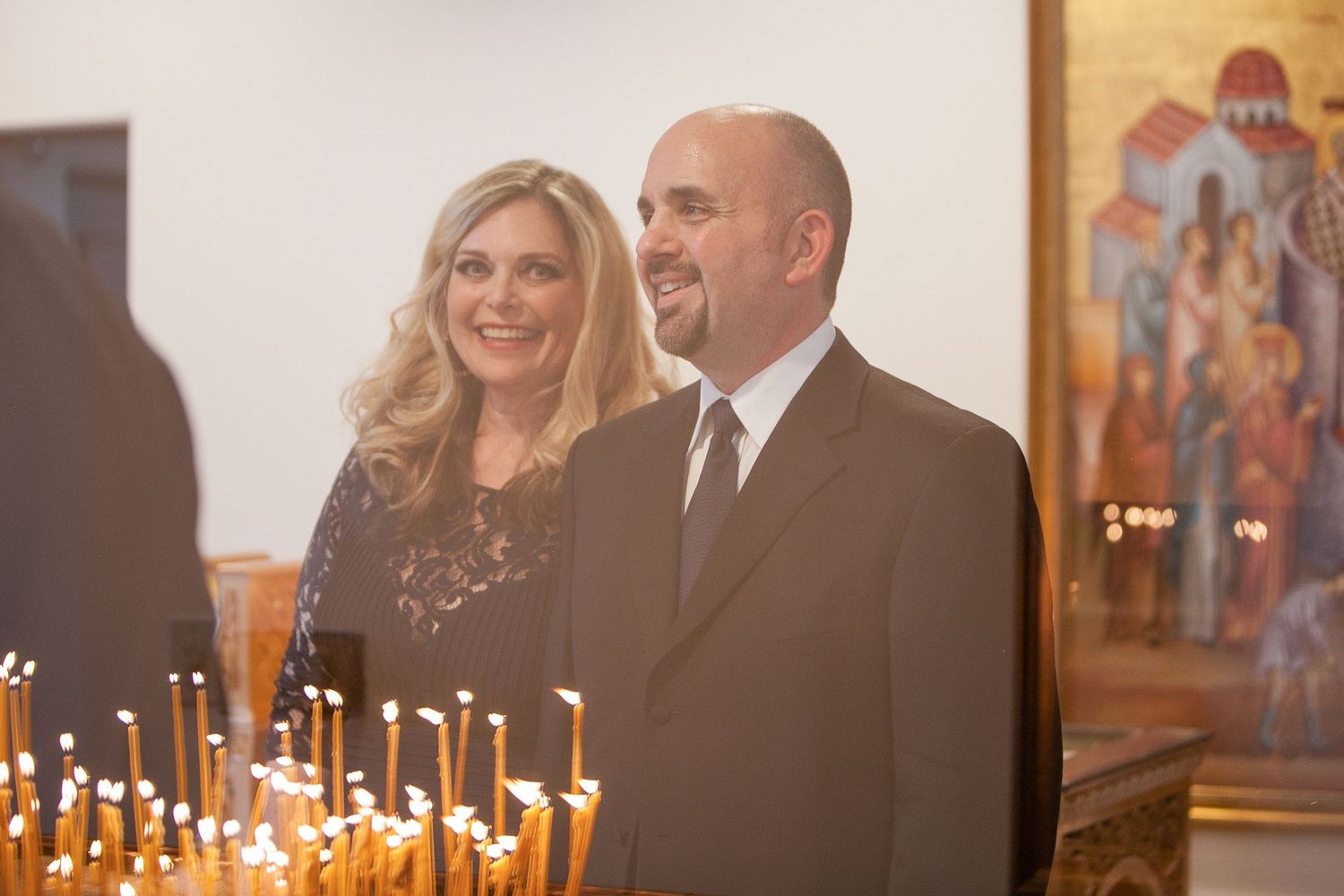 Couple smiling in Orthodox church with candles and religious icons in background.