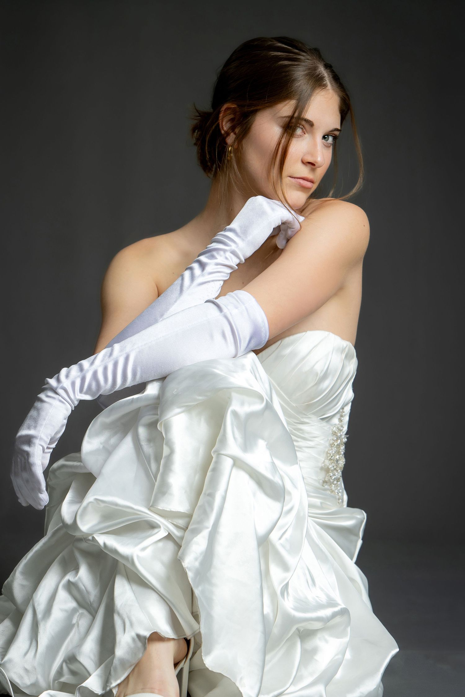 Elegant bride in white strapless satin gown and long white gloves posing gracefully against gray background.