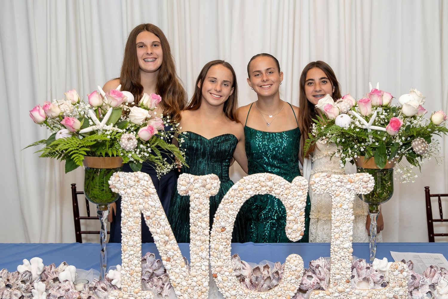 Four girls in green dresses pose behind decorative NCT letters with pink floral arrangements at a formal event.