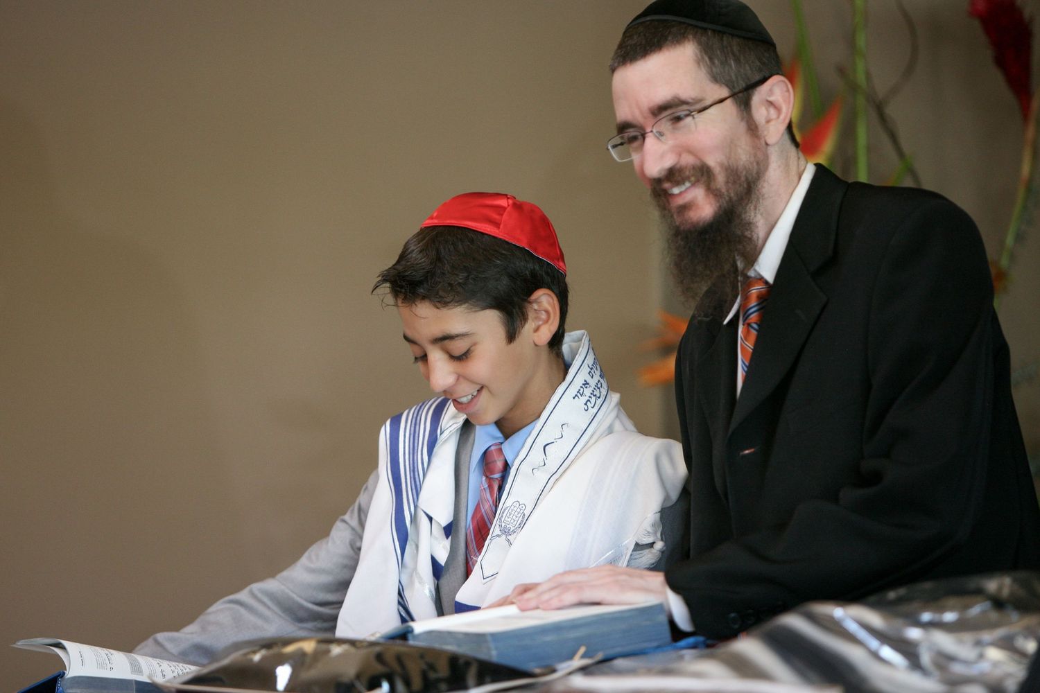 Young boy in kippah and tallit reads Torah at Bar Mitzvah ceremony with rabbi mentor smiling beside him.