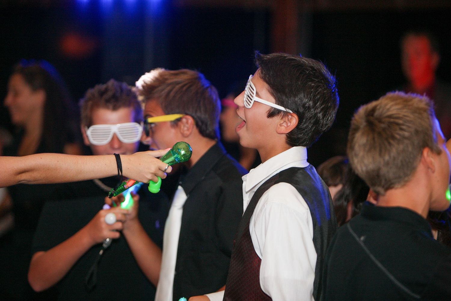 Teens wearing glow-in-the-dark glasses and neon accessories dance at a lively party event.