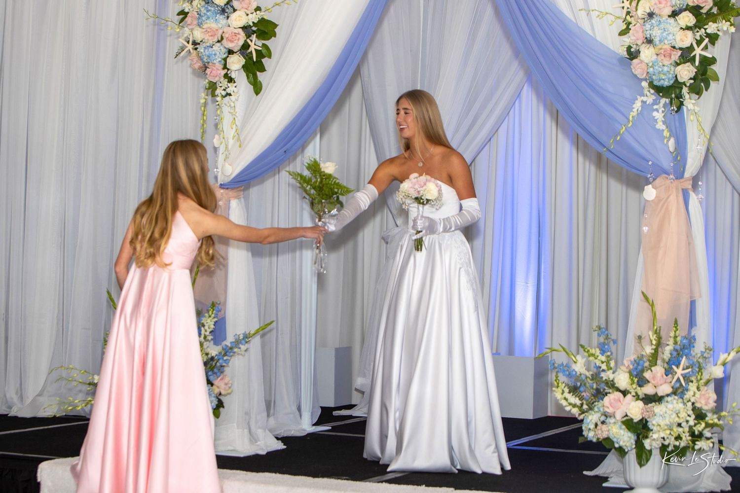 A bride in a white gown receives a bouquet from a girl in pink at an elegant floral wedding ceremony backdrop.