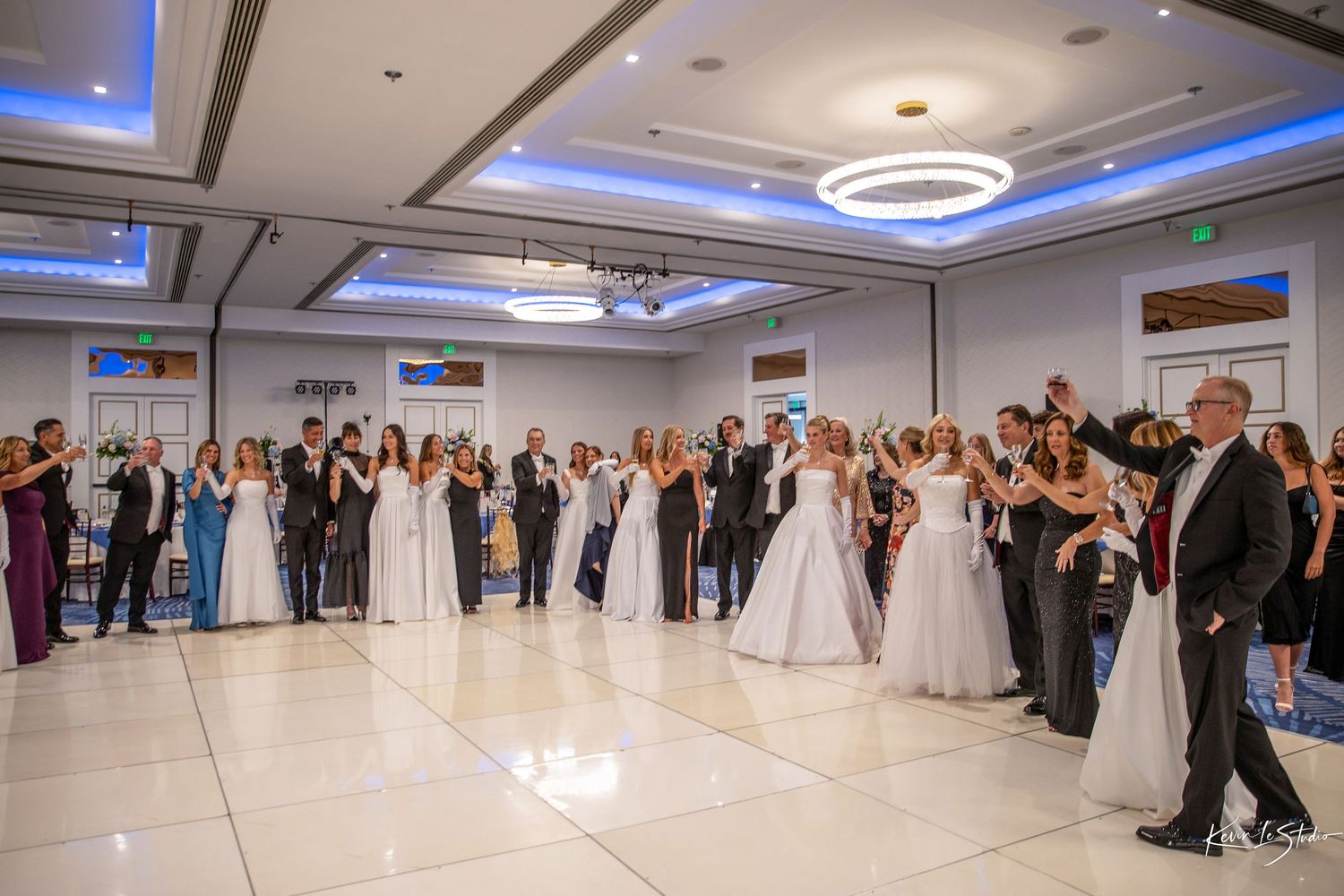 Elegant wedding reception with guests in formal attire gathered on a white dance floor in a decorated ballroom.