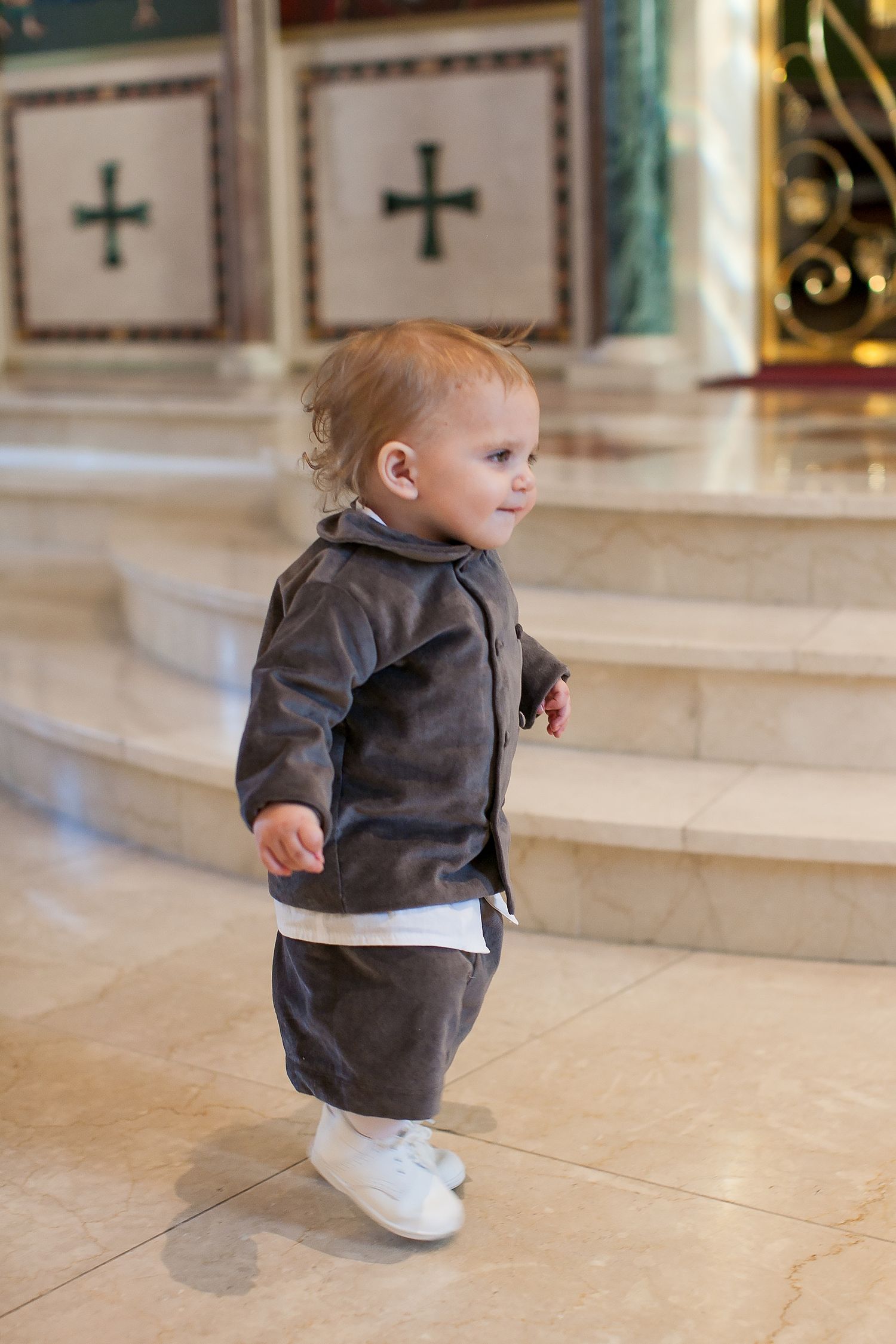 Toddler boy in gray velvet suit walking inside a church with marble altar steps.