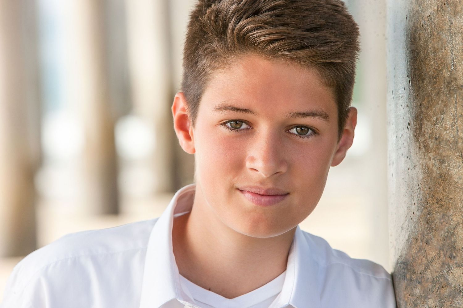 Smiling young boy with brown hair wearing a white shirt, posing outdoors near stone columns.