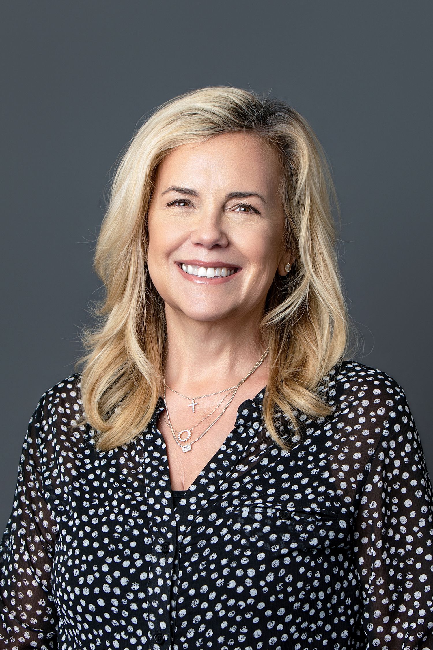 Smiling blonde woman in black polka dot blouse wearing layered necklaces, professional headshot on gray background.