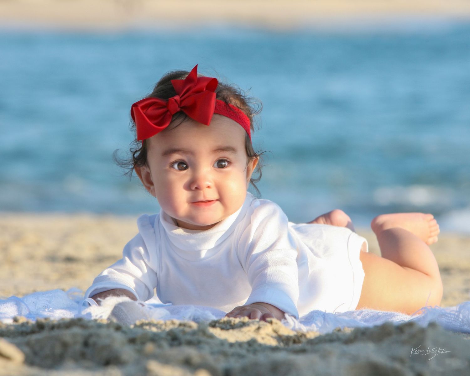 Adorable baby girl with red bow headband lying on sandy beach in white outfit.
