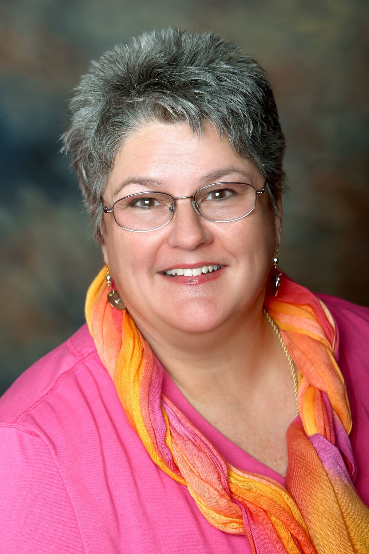 Professional headshot of a smiling older woman with short gray hair, glasses, and colorful orange scarf.