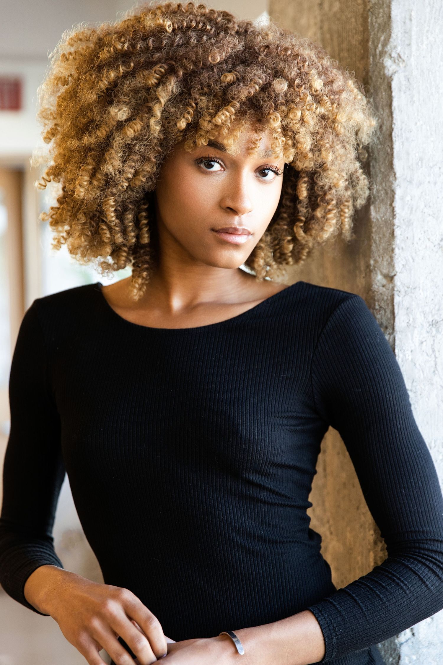 Young Black woman with voluminous blonde curly afro wearing black long-sleeve top, leaning on wall.