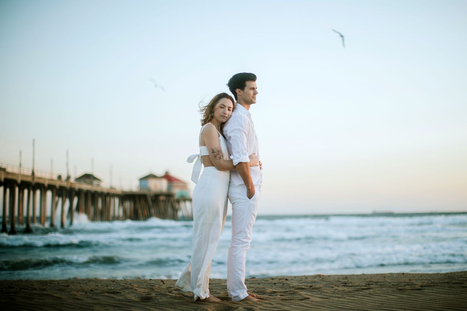 Romantic couple in white outfits embracing on sandy beach near pier at dusk.