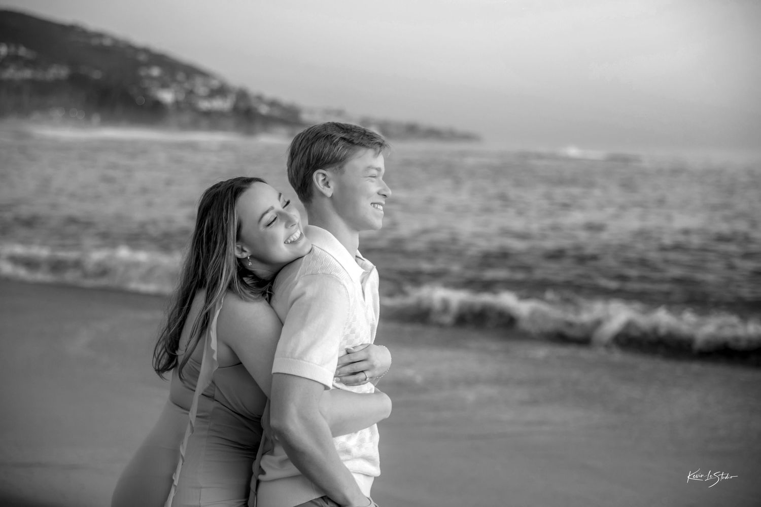 Black and white photo of couple embracing and laughing on beach shoreline.