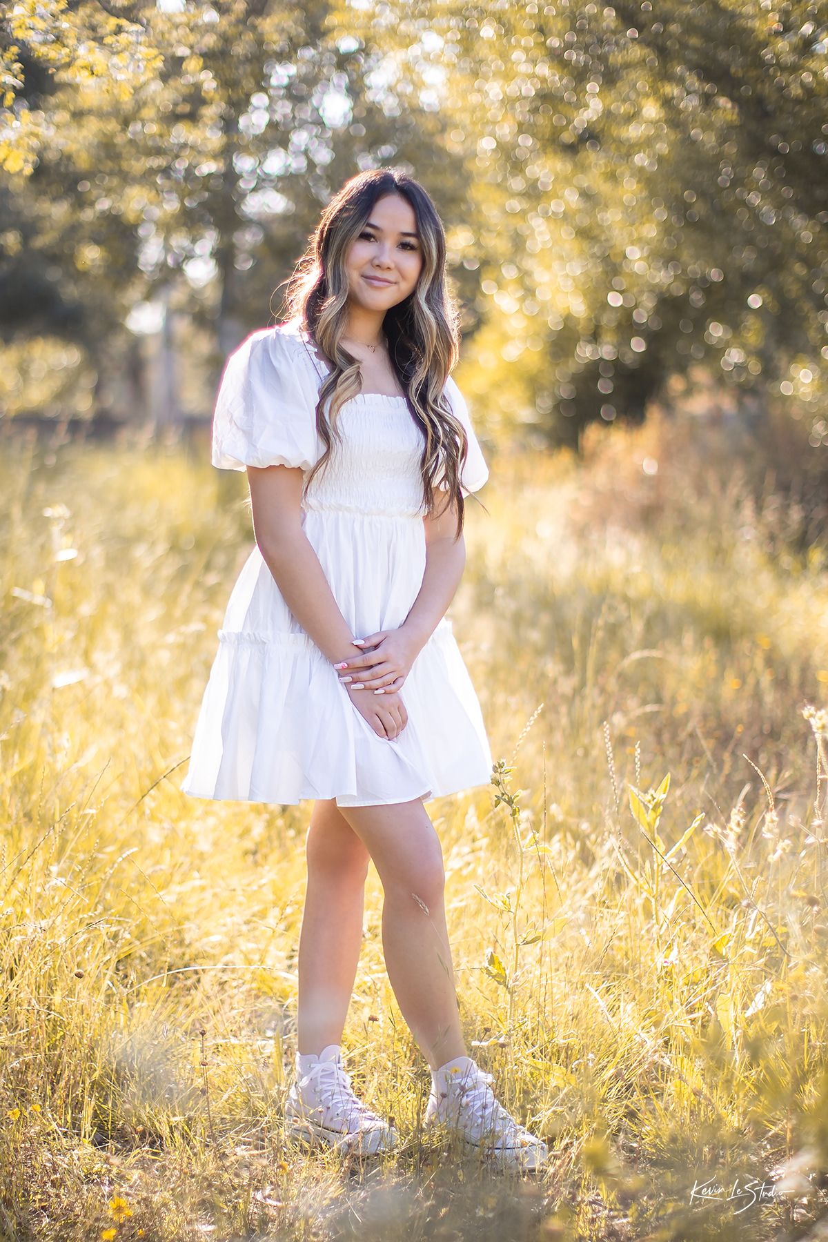 Brunette woman in white tiered mini dress and white sneakers standing in sunny golden field.