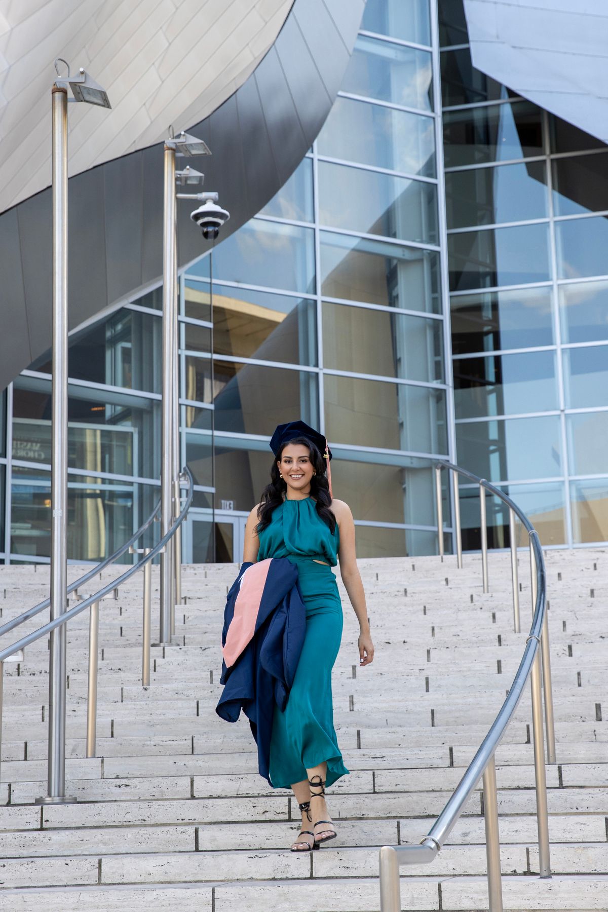 Smiling woman in teal jumpsuit and graduation cap walking down steps outside a modern glass building.