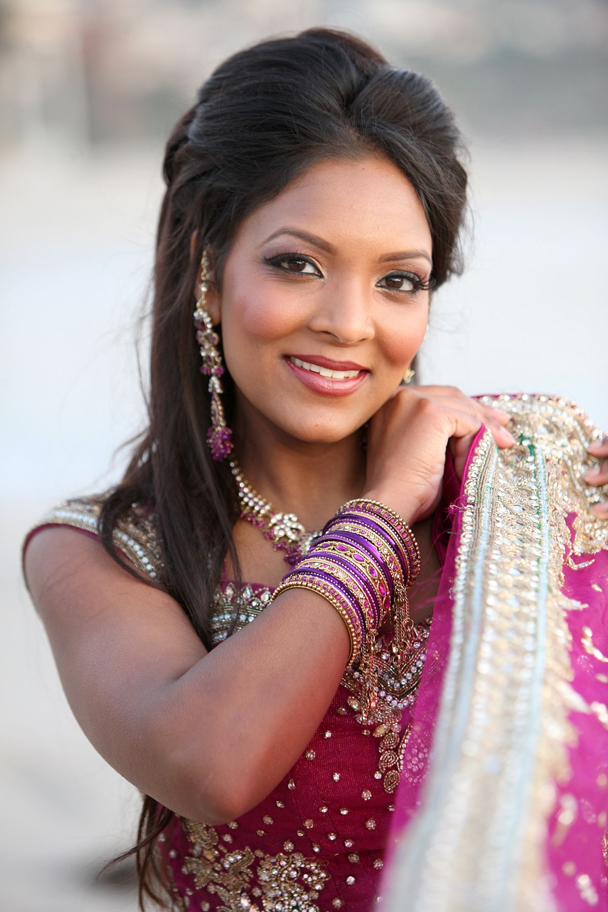 Stunning Indian bride in a magenta lehenga adorned with gold jewelry and colorful bangles smiling outdoors.
