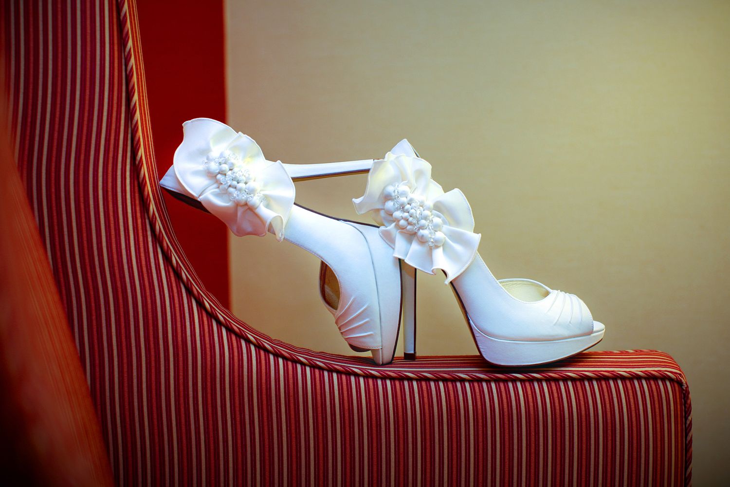 White bridal platform heels with floral bow detail resting on a striped armchair.