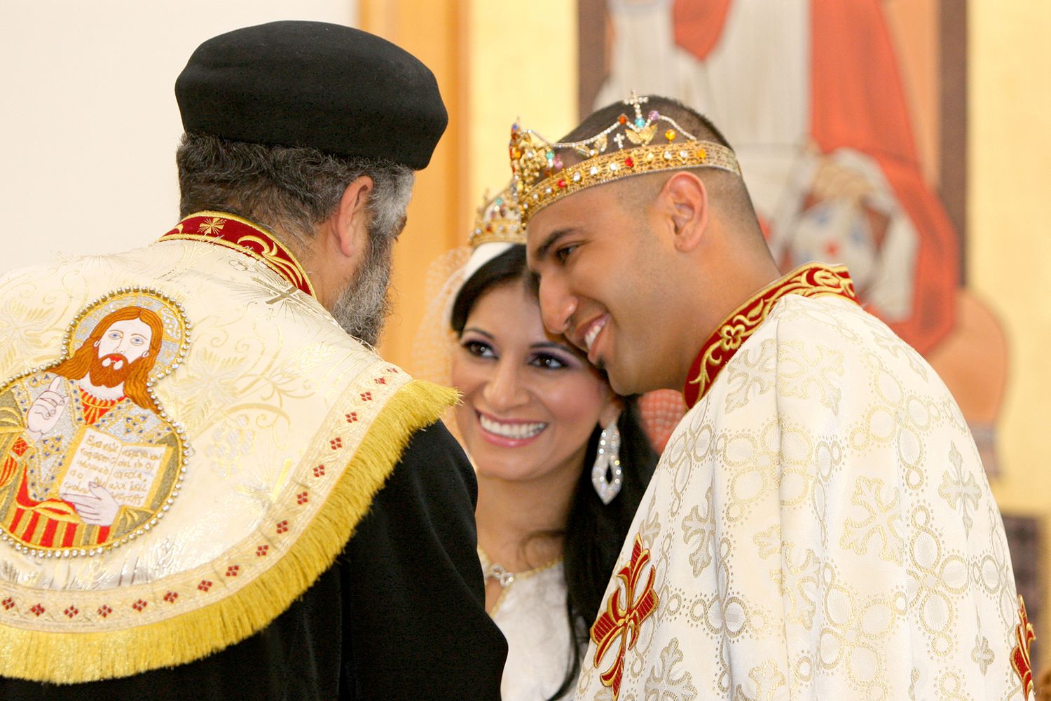 Couple in ornate crowns share an intimate moment with a priest during a traditional Orthodox wedding ceremony.