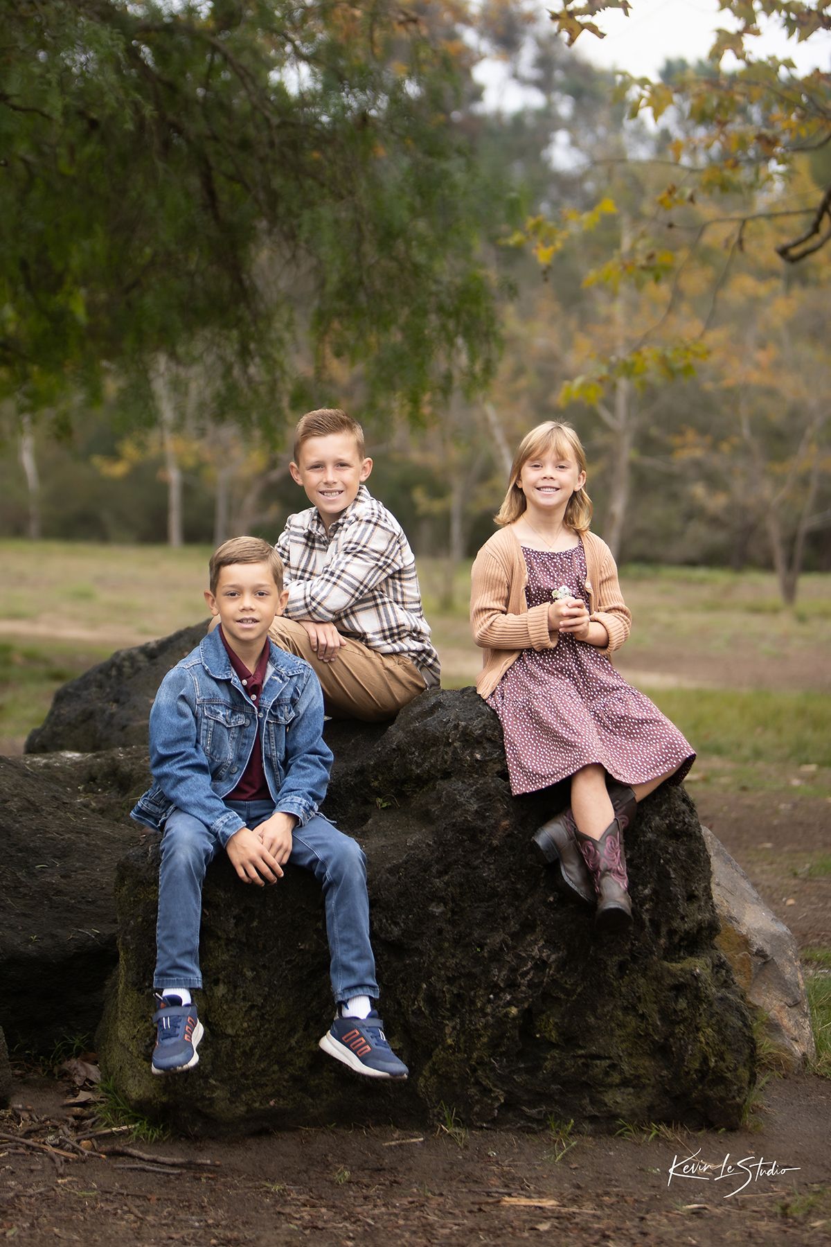 Three children pose on large rocks in a park during fall, wearing casual outfits in an outdoor family portrait.