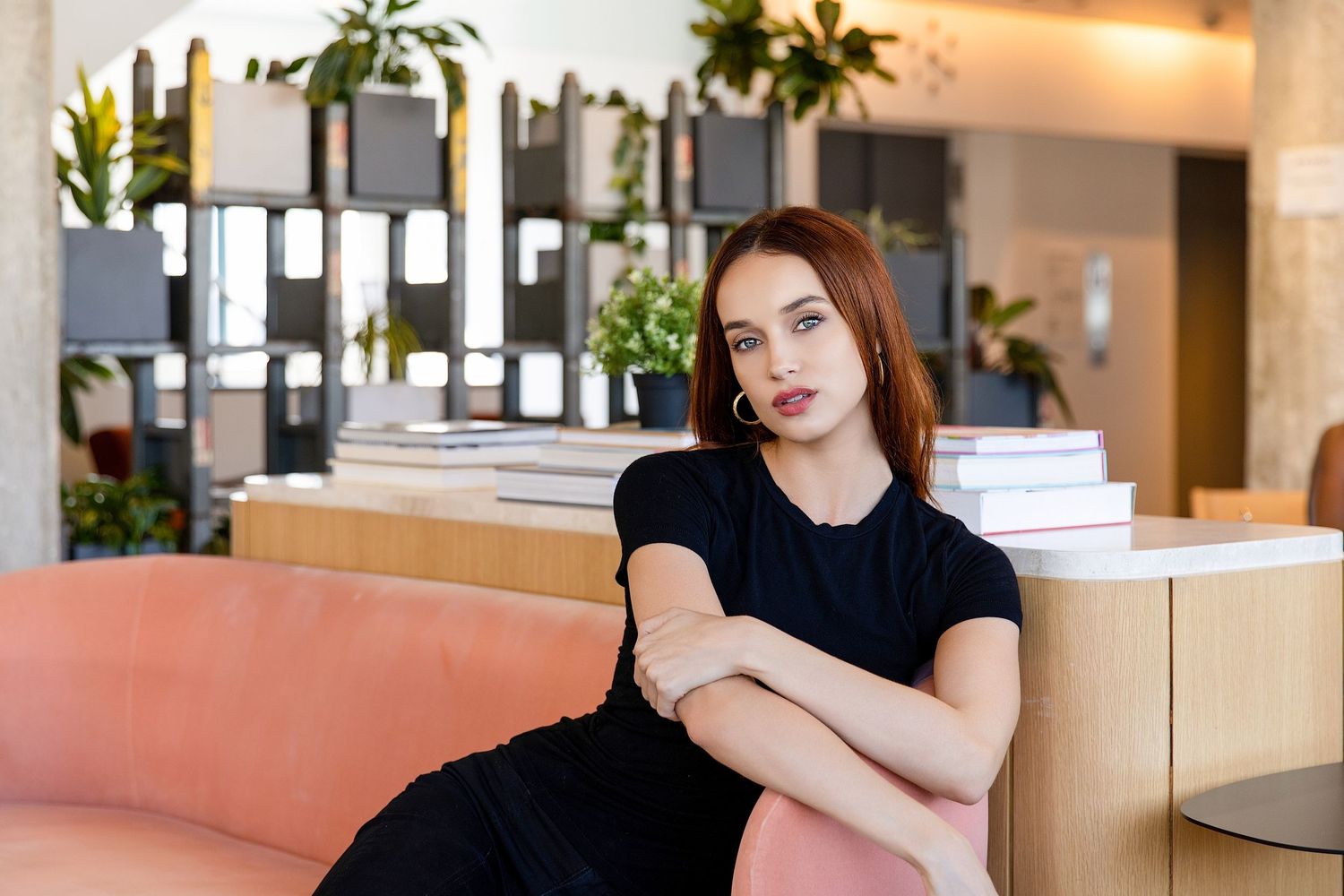 Young woman with auburn hair in black dress sitting cross-armed on pink sofa in modern interior.
