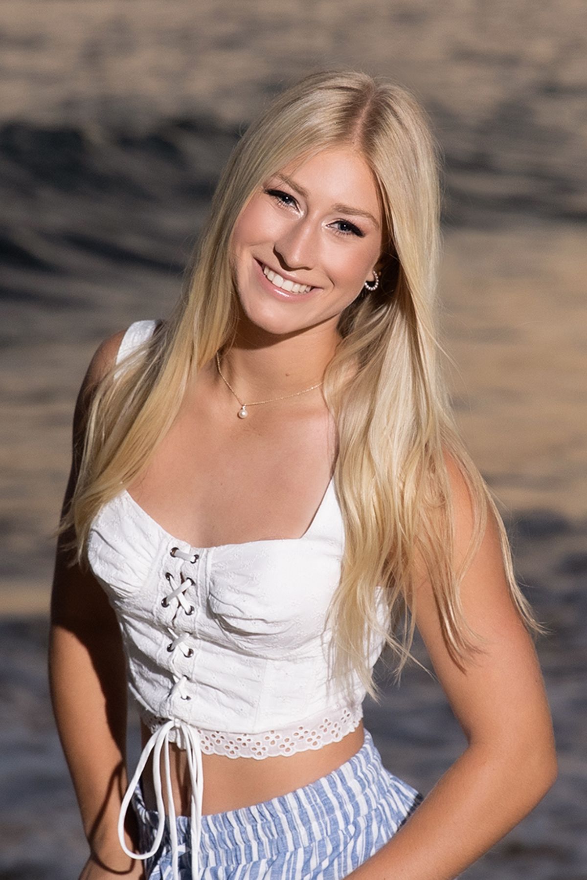 Smiling blonde woman in white corset top and blue striped skirt posing near ocean at golden hour.