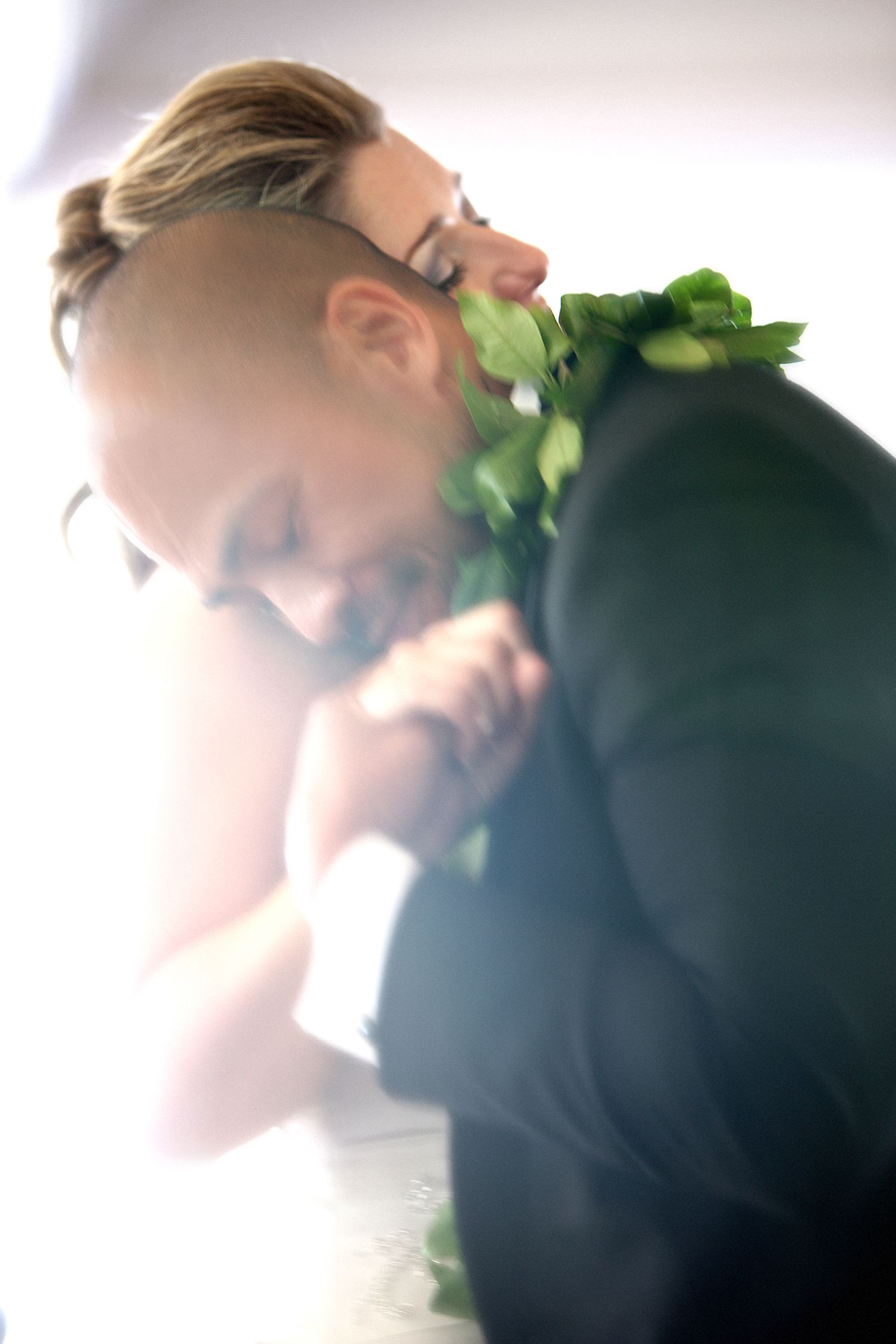 A groom in a black suit with green floral boutonniere tenderly holds hands with his bride in a softly lit wedding photo.