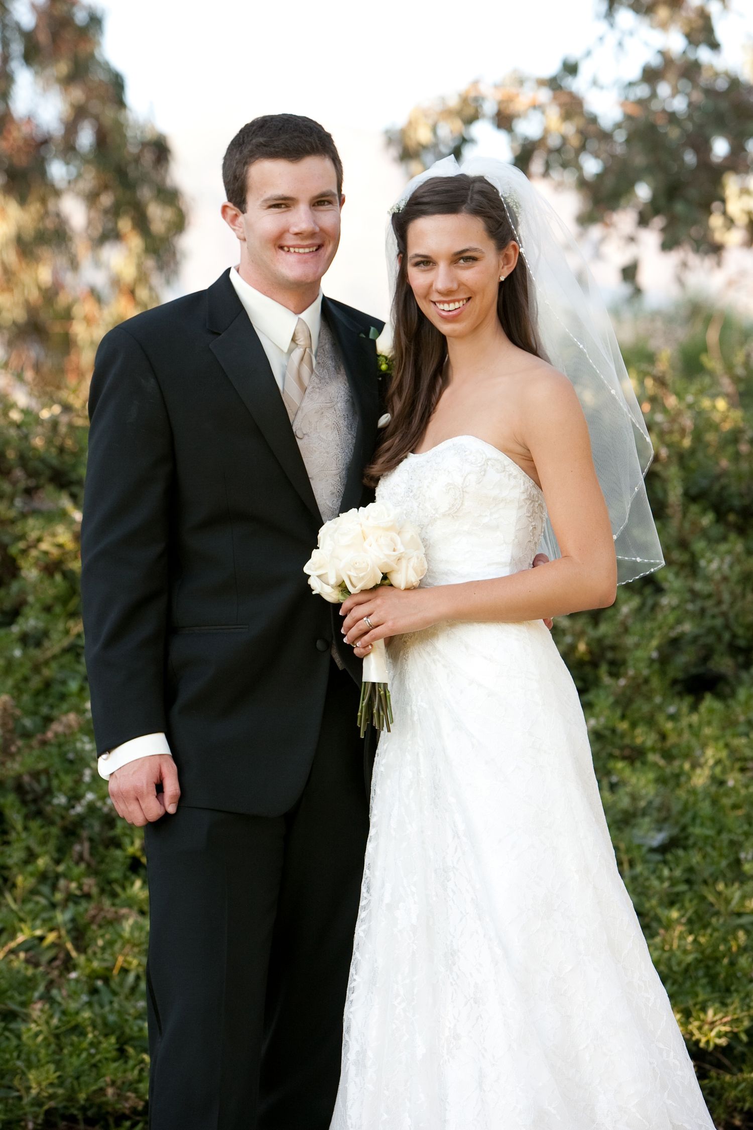 Smiling bride in white strapless gown with veil holding bouquet stands beside groom in black tuxedo outdoors.
